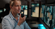 A woman in a blue security shirt speaks into a walkie-talkie while monitoring security footage on desk-mounted screens.