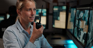 A woman in a blue security shirt speaks into a walkie-talkie while monitoring security footage on desk-mounted screens.