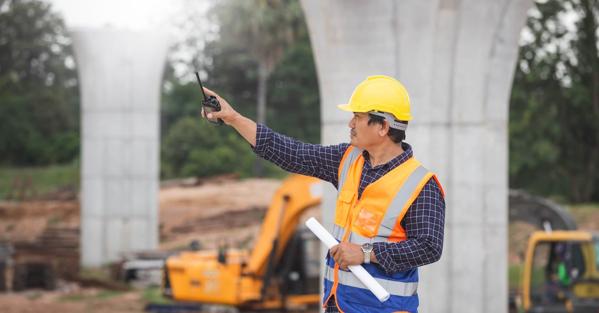 A construction worker pointing in the distance while holding a two-way radio and rolled-up paper, with equipment behind him.