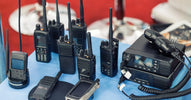 A variety of two-way radio equipment is displayed on a small table with a blue tablecloth at a tech convention.