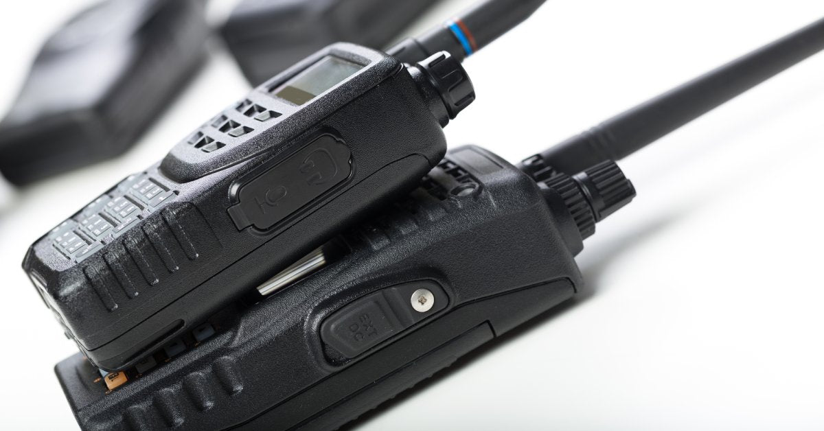 Two different types of two-way radios are stacked on top of each other on a white table with more radios in the background.
