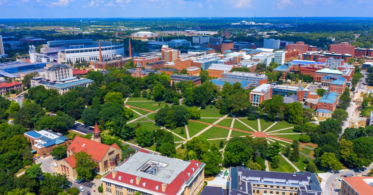 An aerial view of a large college campus shows intersecting paved walkways in a green space with a stadium in the distance.