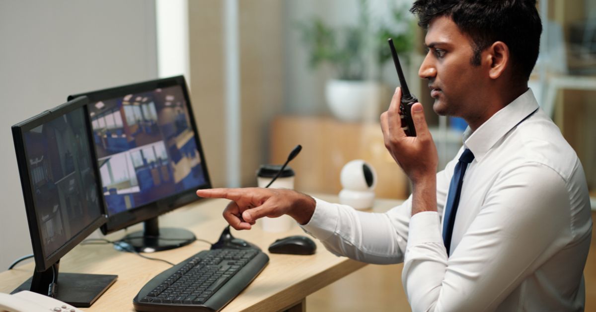 A young male security guard points at a surveillance monitor while speaking into a two-way radio at his desk.
