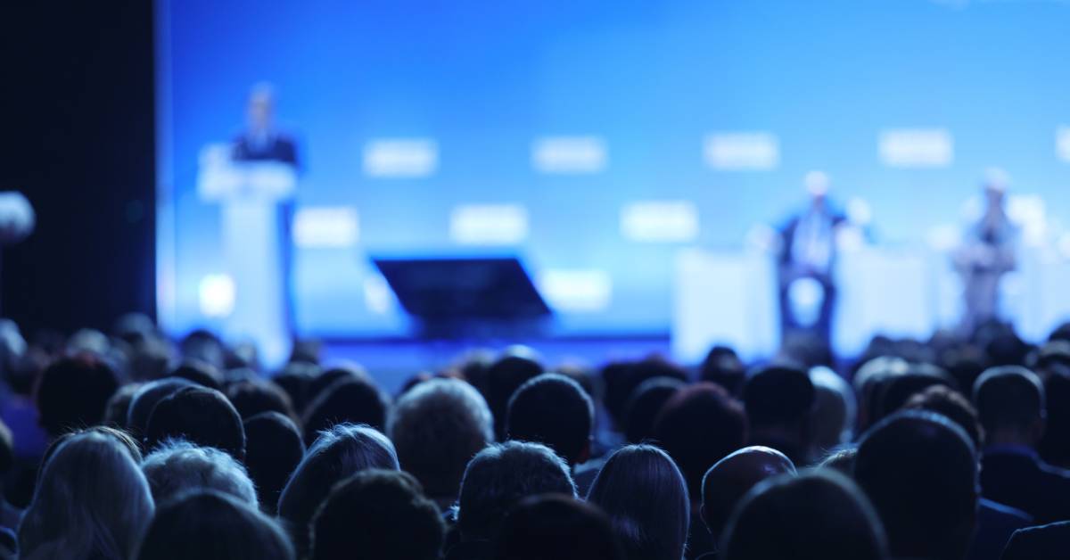 A crowd of people sits facing an out-of-focus stage with blue lighting where a person speaks at a podium.