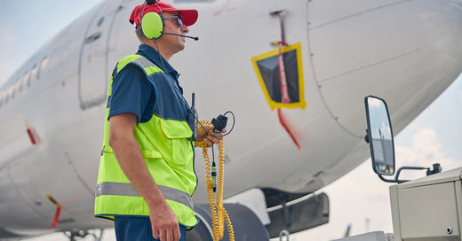 A man wearing a red hat, high-visibility vest, and over the ear headphones, holding a two-way radio in front of a plane.