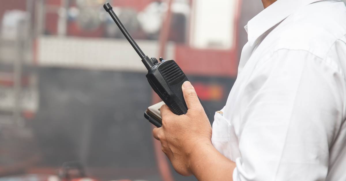 A person wearing a white shirt holds a black two-way radio in front of a blurry firetruck.