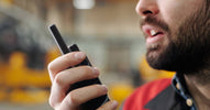 A close-up of a man with a full beard holding and speaking into a portable two-way radio in an industrial setting.