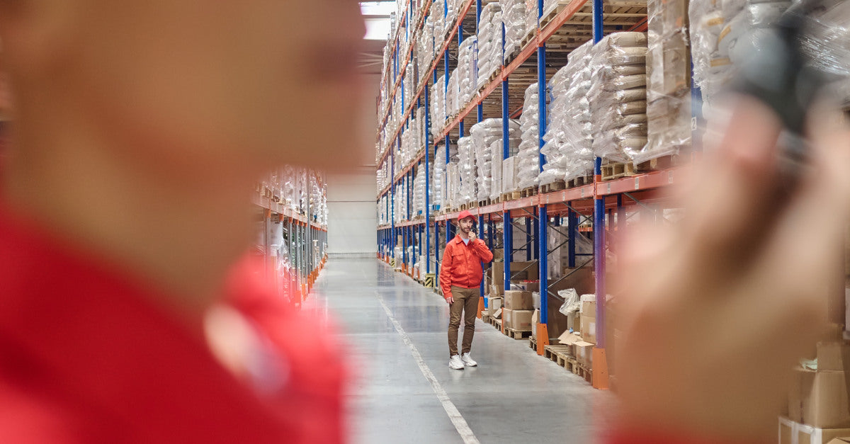 Two warehouse workers wearing red jackets are communicating across the warehouse using two-way radios.