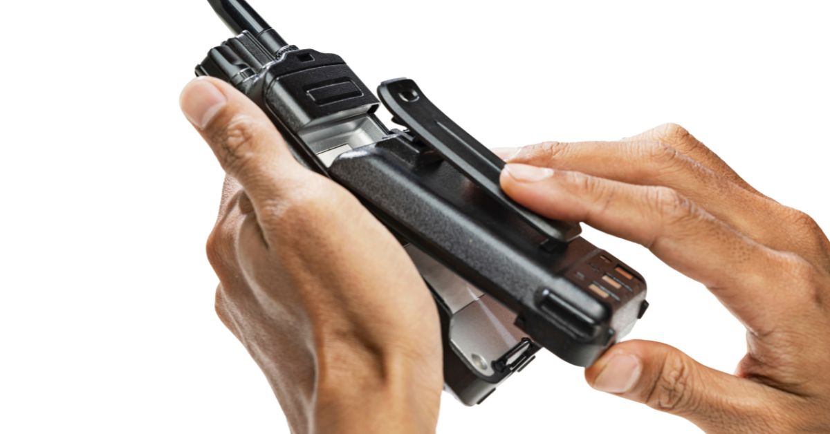 A person holds a black two-way radio and removes its battery, with their hands and the device against a white background.